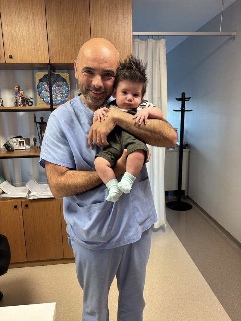 Male doctor in blue scrubs holding a baby in a medical clinic examination room