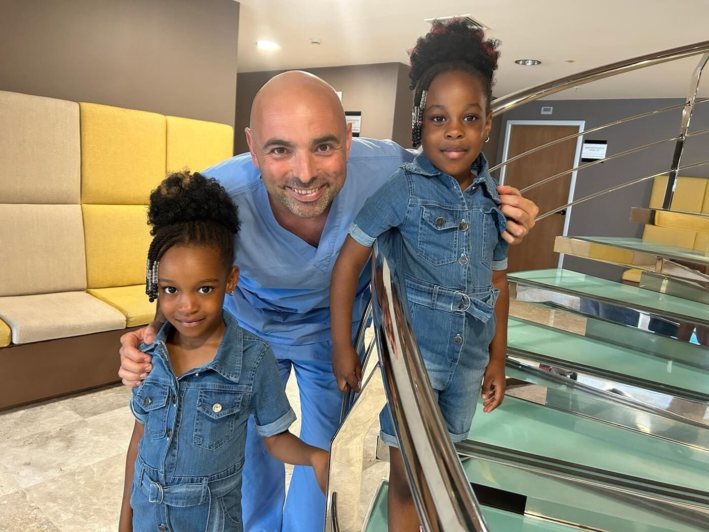 Smiling doctor in blue scrubs with two young girls in denim outfits standing on a staircase indoors