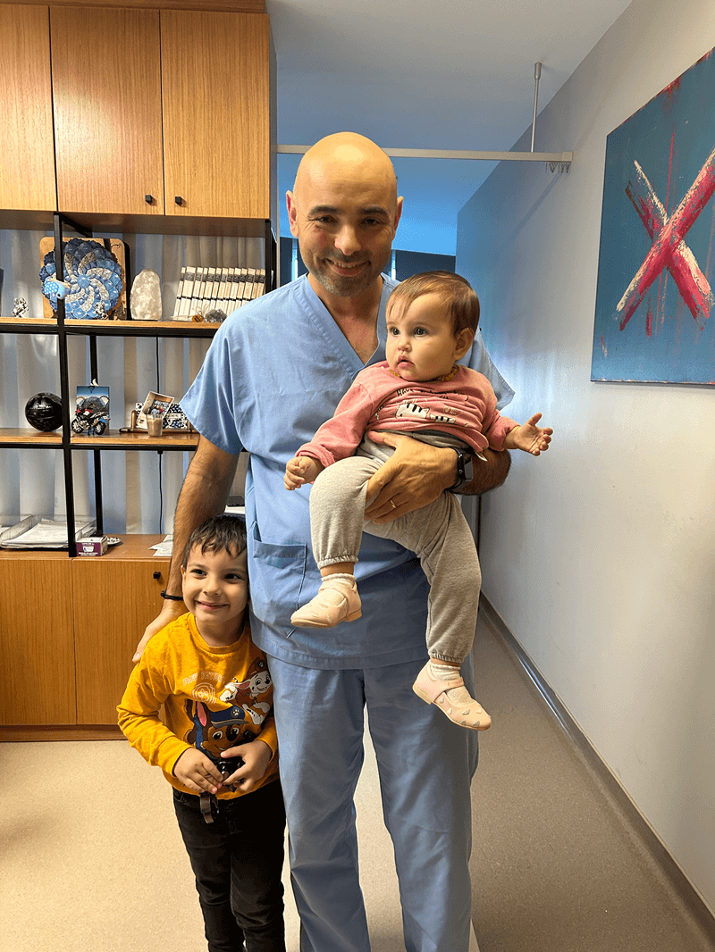 Doctor in blue scrubs holding a baby with a smiling child beside him in a modern clinic interior